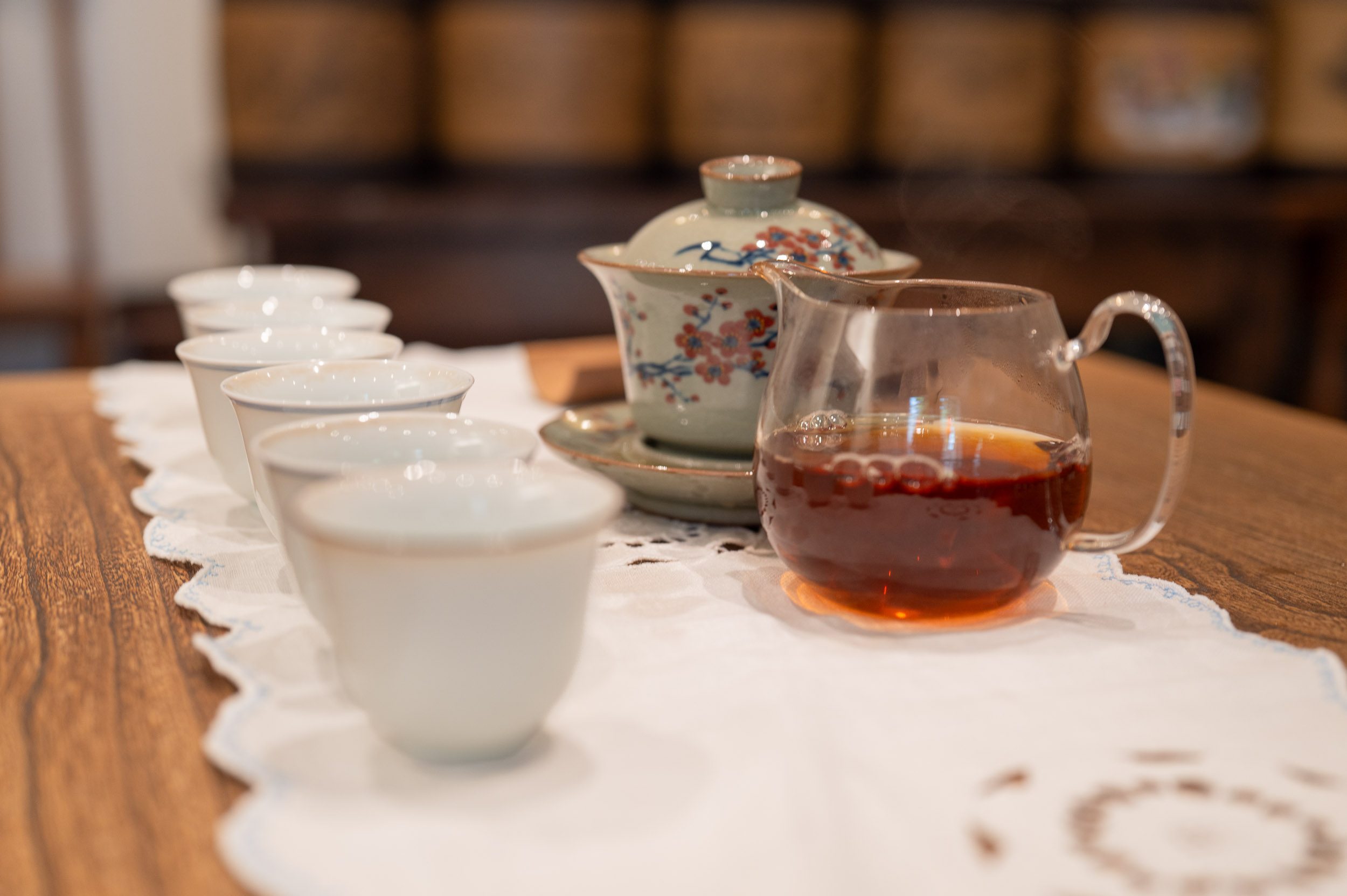 Un gaiwan en céladon et une pitchette en verre remplie de thé rouge fumant, posés sur un chemin de table en dentelle à la boutique L'origine du thé